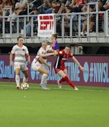 A competitive soccer match is underway with players vying for the ball near the sideline. The players wear different uniforms, indicating they are from opposing teams. The audience is visible in the stadium seating above, and there are visible advertisements and hashtags on the LED boards along the field.
