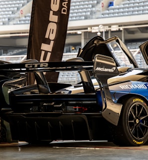 A high-performance sports car with a sleek design is parked in a garage setting. The vehicle features prominent sponsorship logos, including McLaren and Richard Mille. The garage door is open, revealing a glimpse of stadium seating in the background.