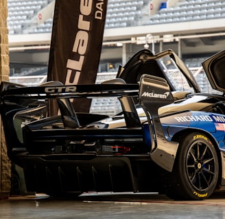 A high-performance sports car with a sleek design is parked in a garage setting. The vehicle features prominent sponsorship logos, including McLaren and Richard Mille. The garage door is open, revealing a glimpse of stadium seating in the background.