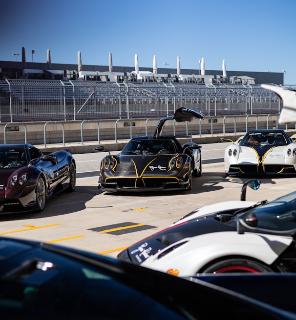 A group of high-end sports cars parked at a racetrack, with one black car prominently in front with its doors open upwards. The background features a racing facility, complete with grandstands and a metal fence. The sky is clear and blue, contributing to the overall sleek and futuristic ambiance.
