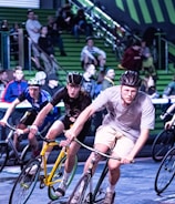 A group of cyclists competes in an indoor race, navigating a track with intense focus. They wear helmets and sport attire, with a crowd of spectators watching intently from the green bleachers in the background.